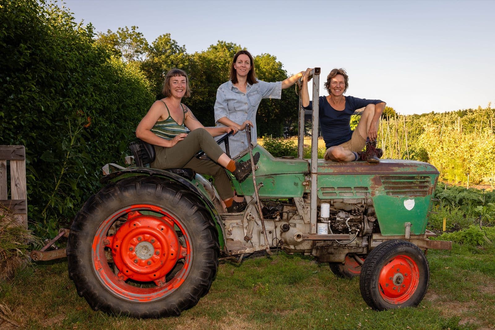 Hella, Tolke en Anne zitten lachend op een oude groene tractor, met de tuin en boomgaard in de avondzon erachter